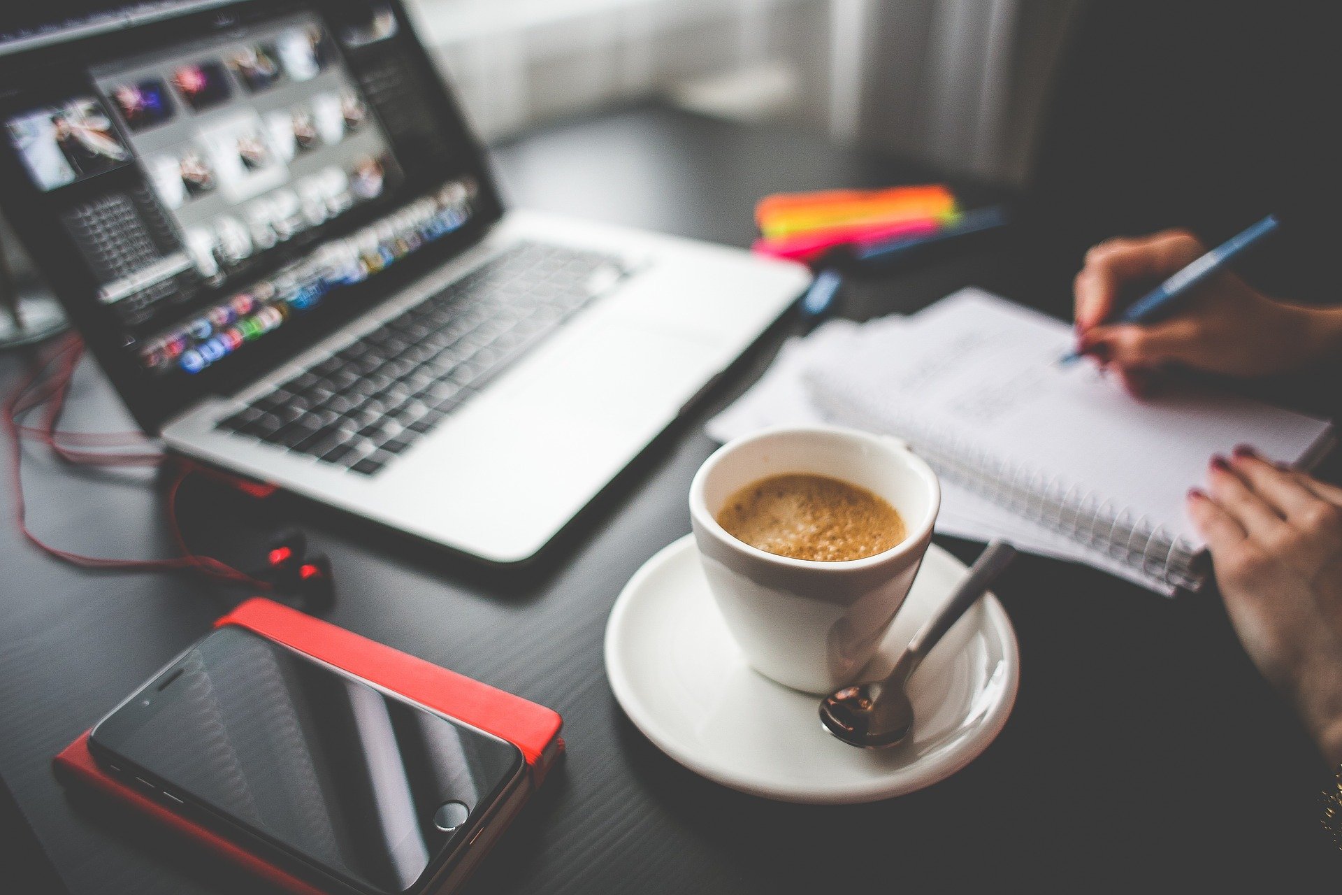 A cup of coffee, phone, and laptop on a table with a person writing in a notebook in the background.