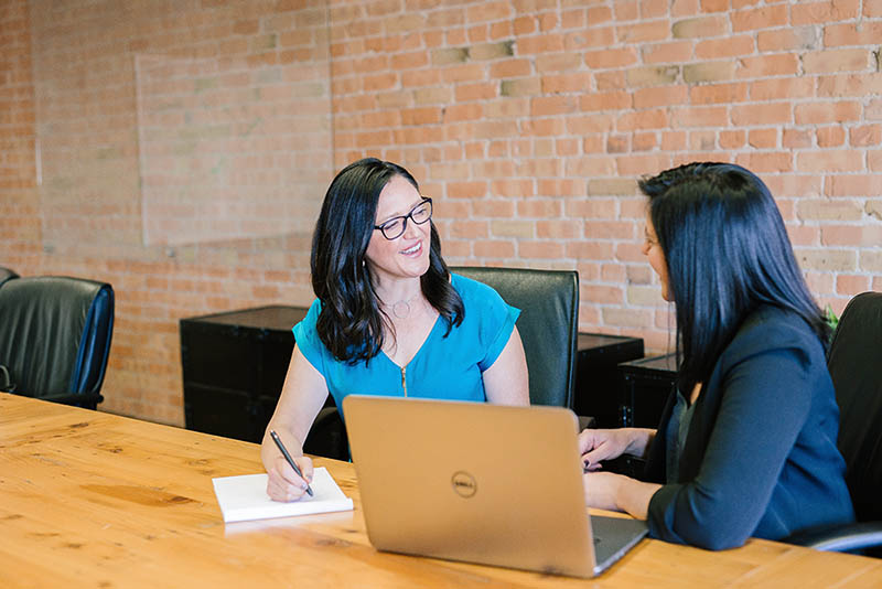 Two women working and talking at a table. One is taking notes on paper and the other is using a laptop.