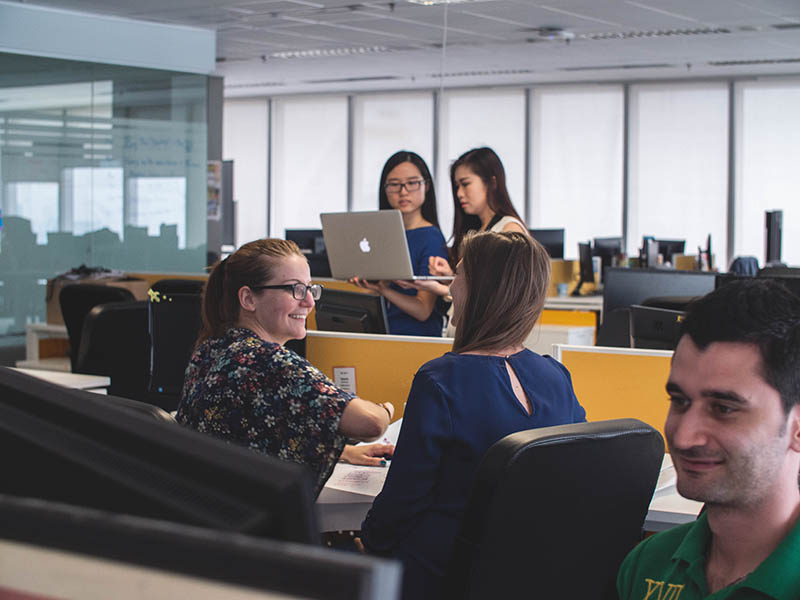 a group of male and female students talking and looking at a laptop in a library study area